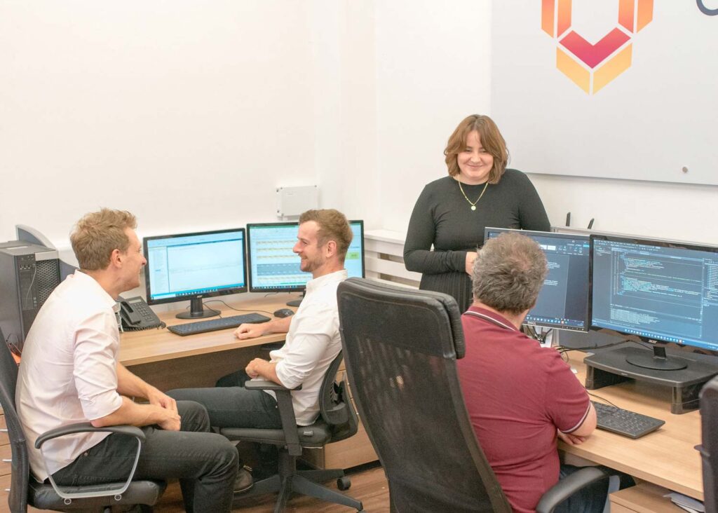 4 people sitting around their computers at the Objective Software development company offices. A girl wearing a black dress, 3 people wearing shirts smiling